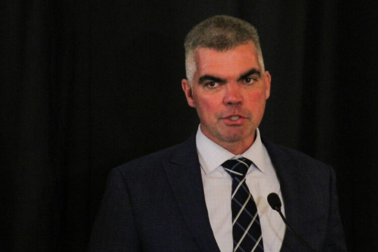 Bob Morris stands in front of a dark blue curtain, wearing a suit and tie. He is a White man with a grey buzzcut that is white at the temples.