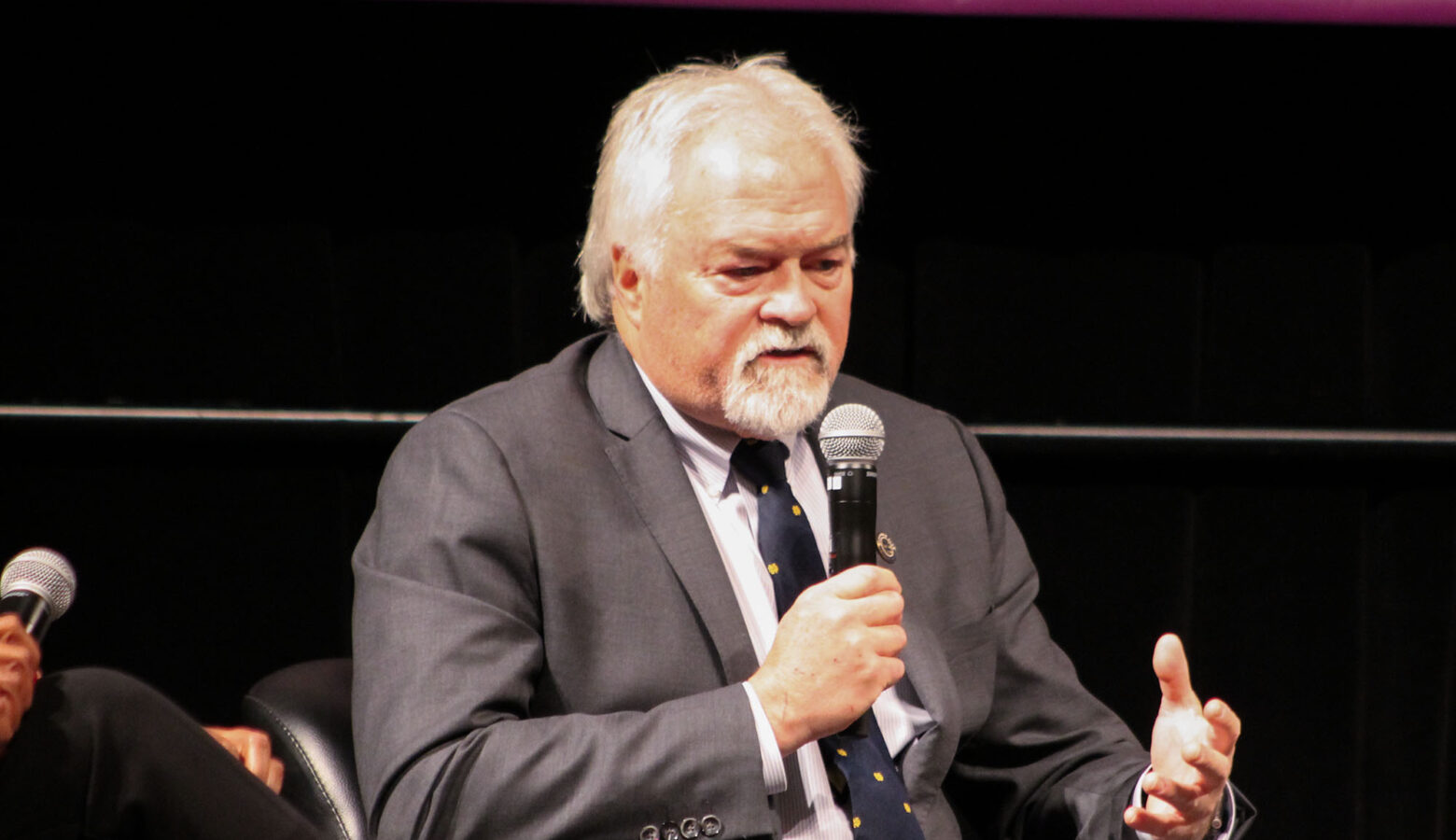 Representative Brad Barrett speaks into a microphone. He's a White man with thinning white hair and a goatee wearing a suit and tie.