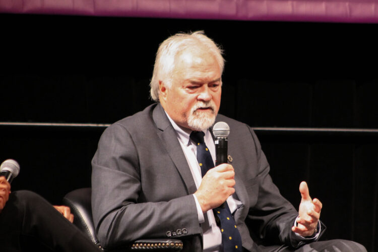 Representative Brad Barrett speaks into a microphone. He's a White man with thinning white hair and a goatee wearing a suit and tie.