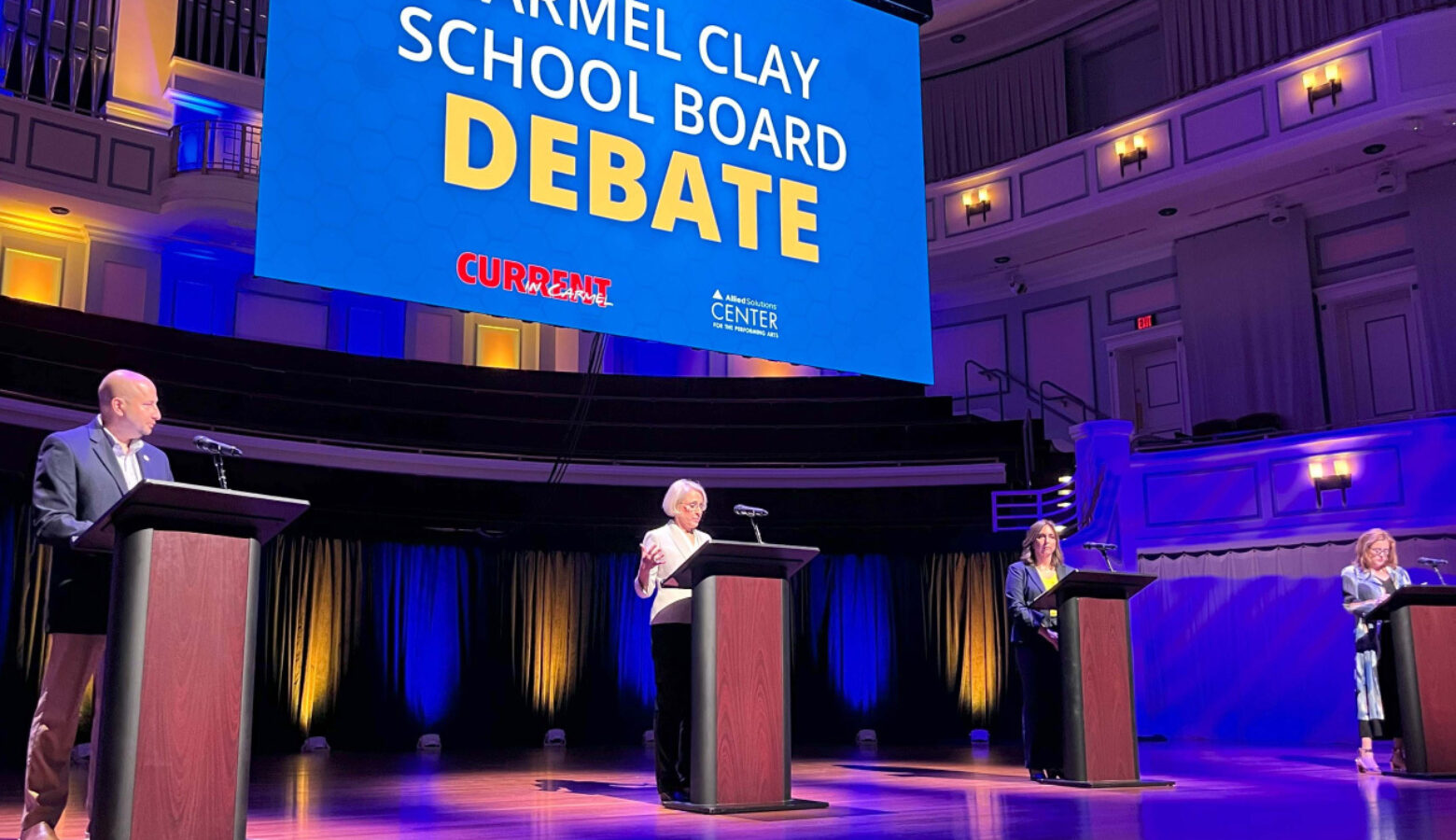 Candidates stand behind podiums on a stage. A sign that reads "Carmel Clay School Board Debate" hangs above them.