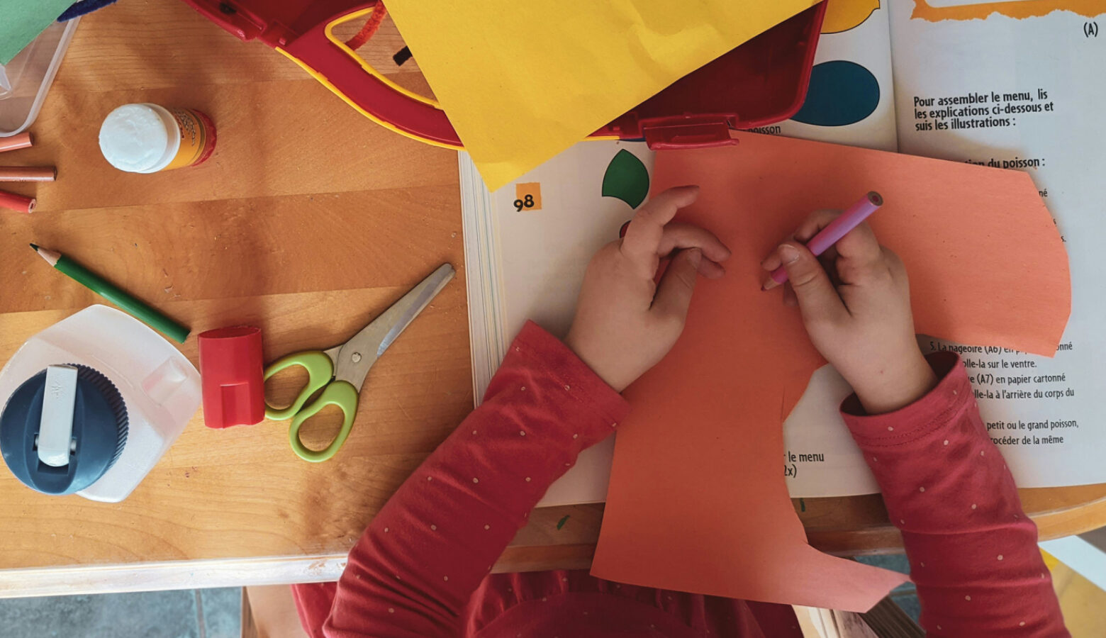 A desk with art supplies is seen from above, as a child with a red shit draws on orange construction paper.