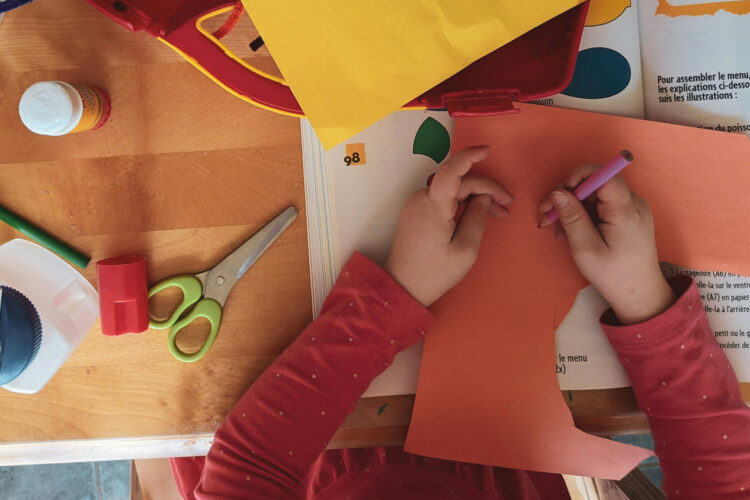 A desk with art supplies is seen from above, as a child with a red shit draws on orange construction paper.