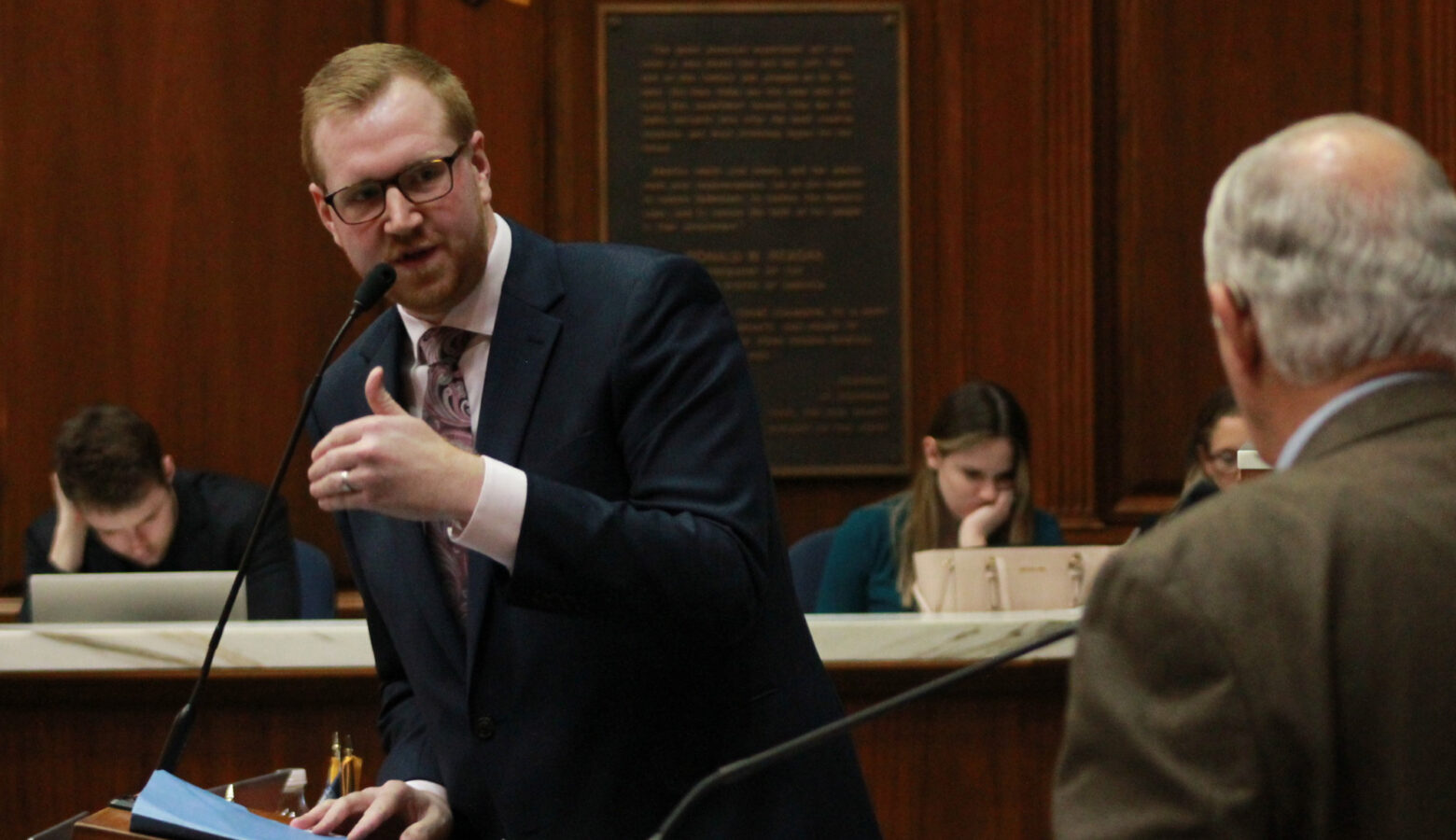 Representative Ethan Manning wearing a navy blue suit with a white shirt and a paisley patterned tie speaking into a microphone while debating another lawmaker while on the House floor.