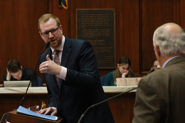 Representative Ethan Manning wearing a navy blue suit with a white shirt and a paisley patterned tie speaking into a microphone while debating another lawmaker while on the House floor.