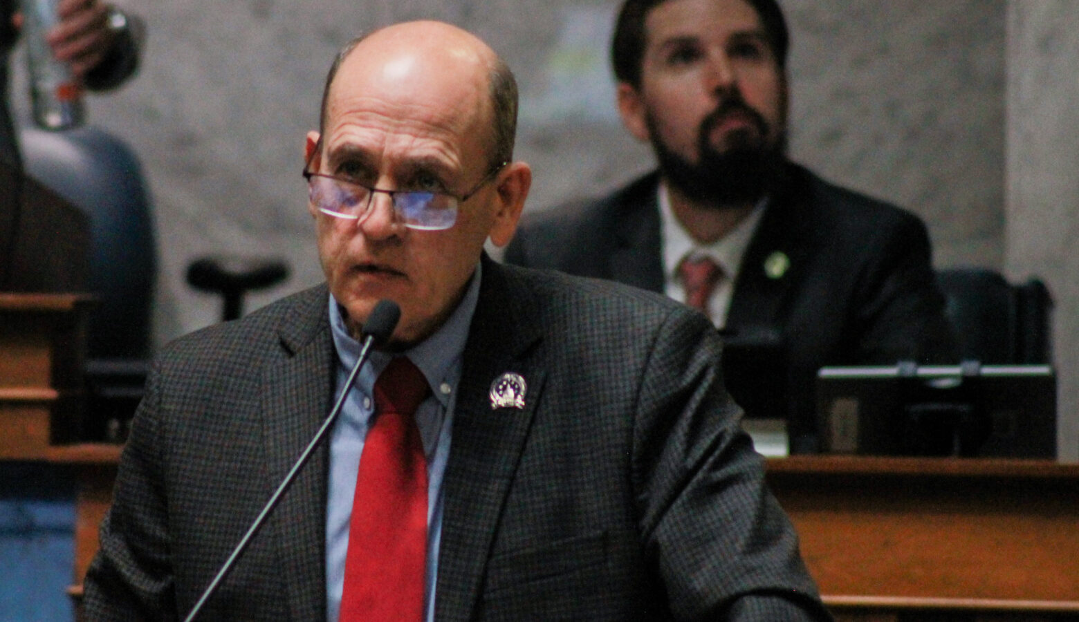 Gary Byrne speaks on the Senate floor. Byrne is a White man, mostly bald. He is wearing glasses and a dark suit.