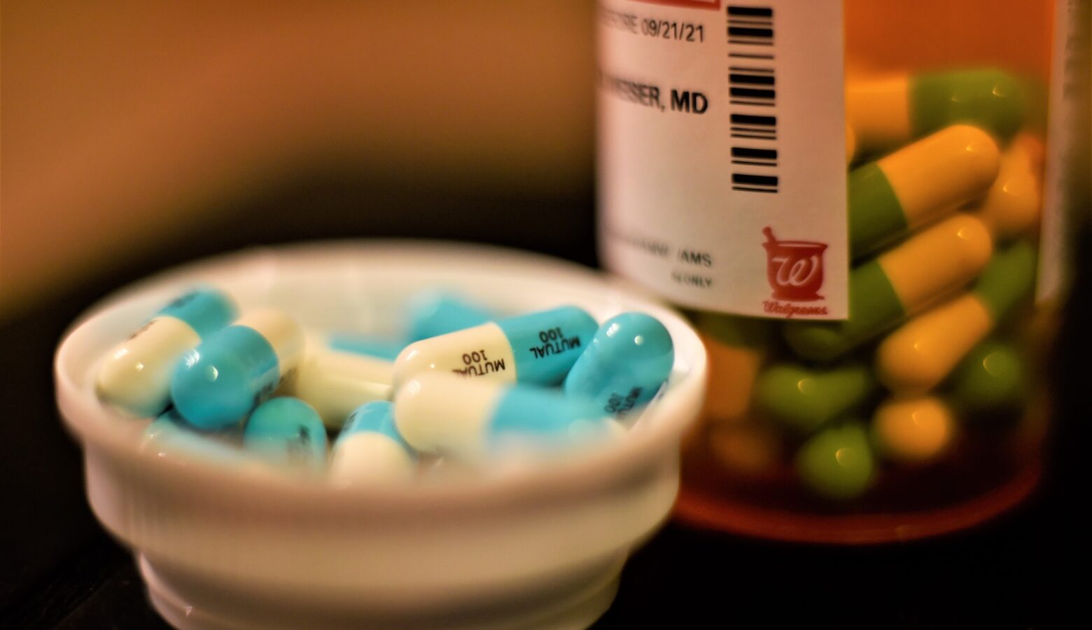 An orange prescription pill bottle sits on a dark surface. White and light blue pill capsules have been poured inside the lid, which sits next to the bottle filled with the same pills.