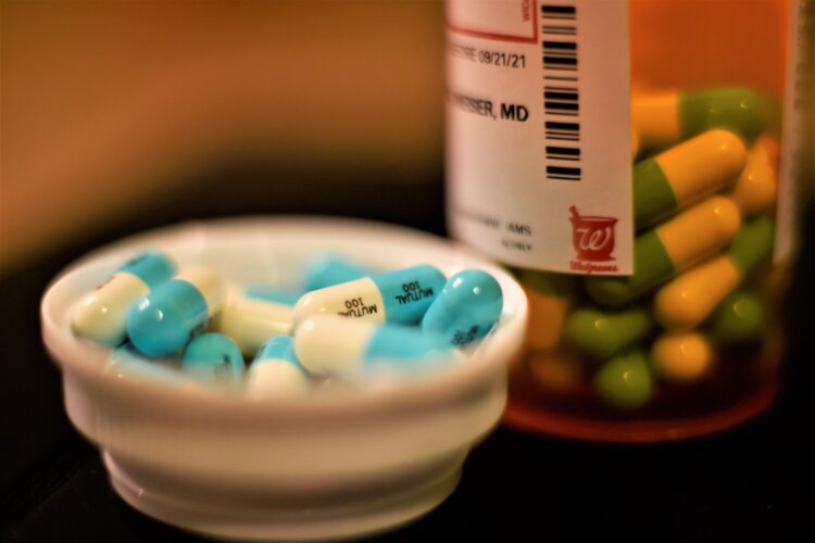 An orange prescription pill bottle sits on a dark surface. White and light blue pill capsules have been poured inside the lid, which sits next to the bottle filled with the same pills.