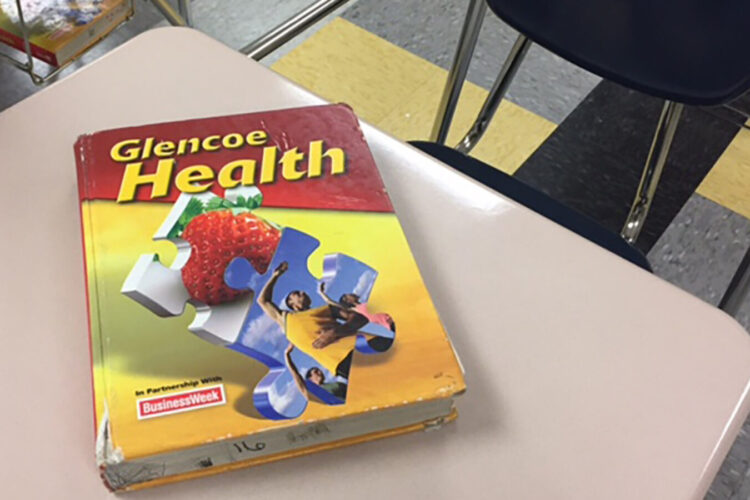 A red and yellow textbook titled "Glencoe Health" with puzzle pieces on the front is displayed on a desk in a school classroom.