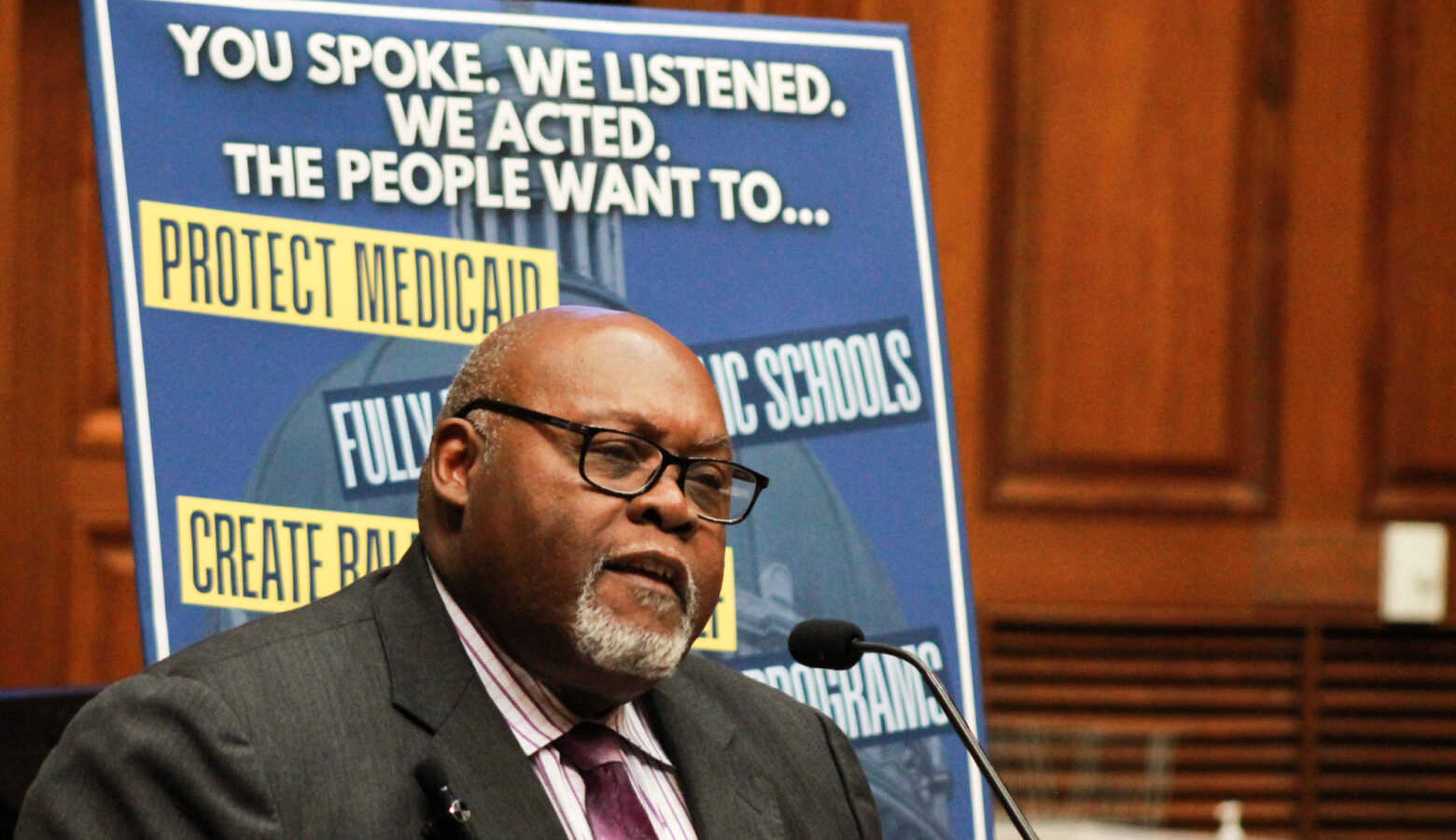 Greg Porter stands in front of a poster that reads "You Spoke. We Listened. We Act. The People Want To..." Porter is a Black man, bald with a white goatee. He is wearing glasses and a suit and tie.