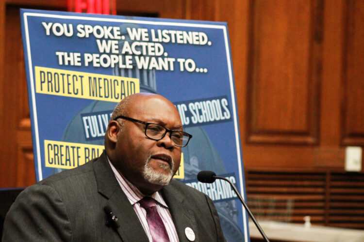 Greg Porter stands in front of a poster that reads "You Spoke. We Listened. We Act. The People Want To..." Porter is a Black man, bald with a white goatee. He is wearing glasses and a suit and tie.