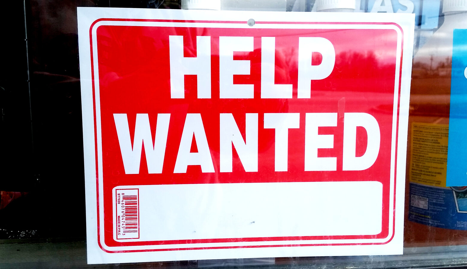 A red sign with a white border reads "HELP WANTED" in capital bold white letters inside of a window.