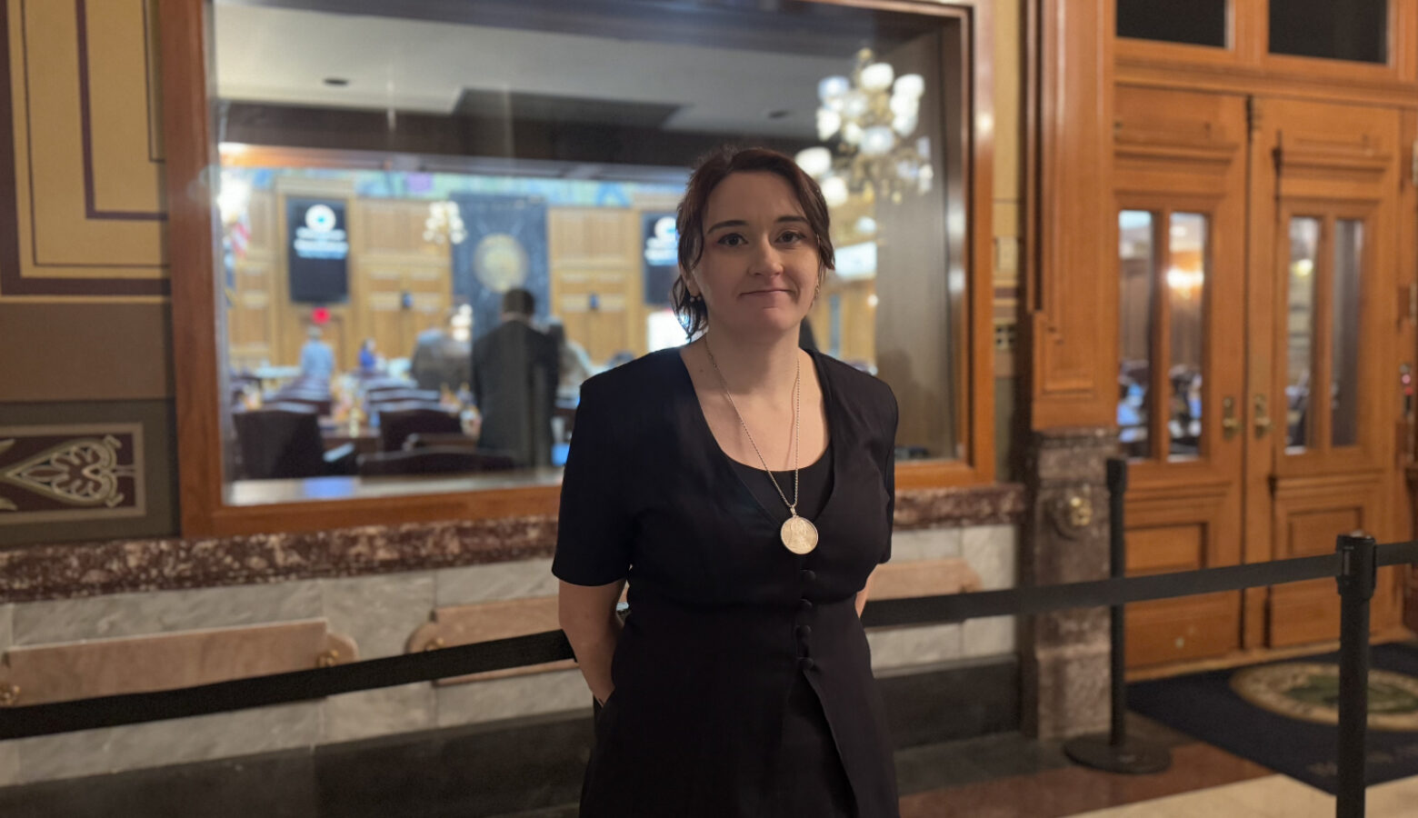 Sarah Conrad stands inside the Statehouse. She's a White woman, wearing a black top and a large gold necklace.