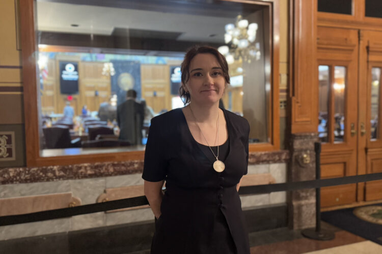 Sarah Conrad stands inside the Statehouse. She's a White woman, wearing a black top and a large gold necklace.