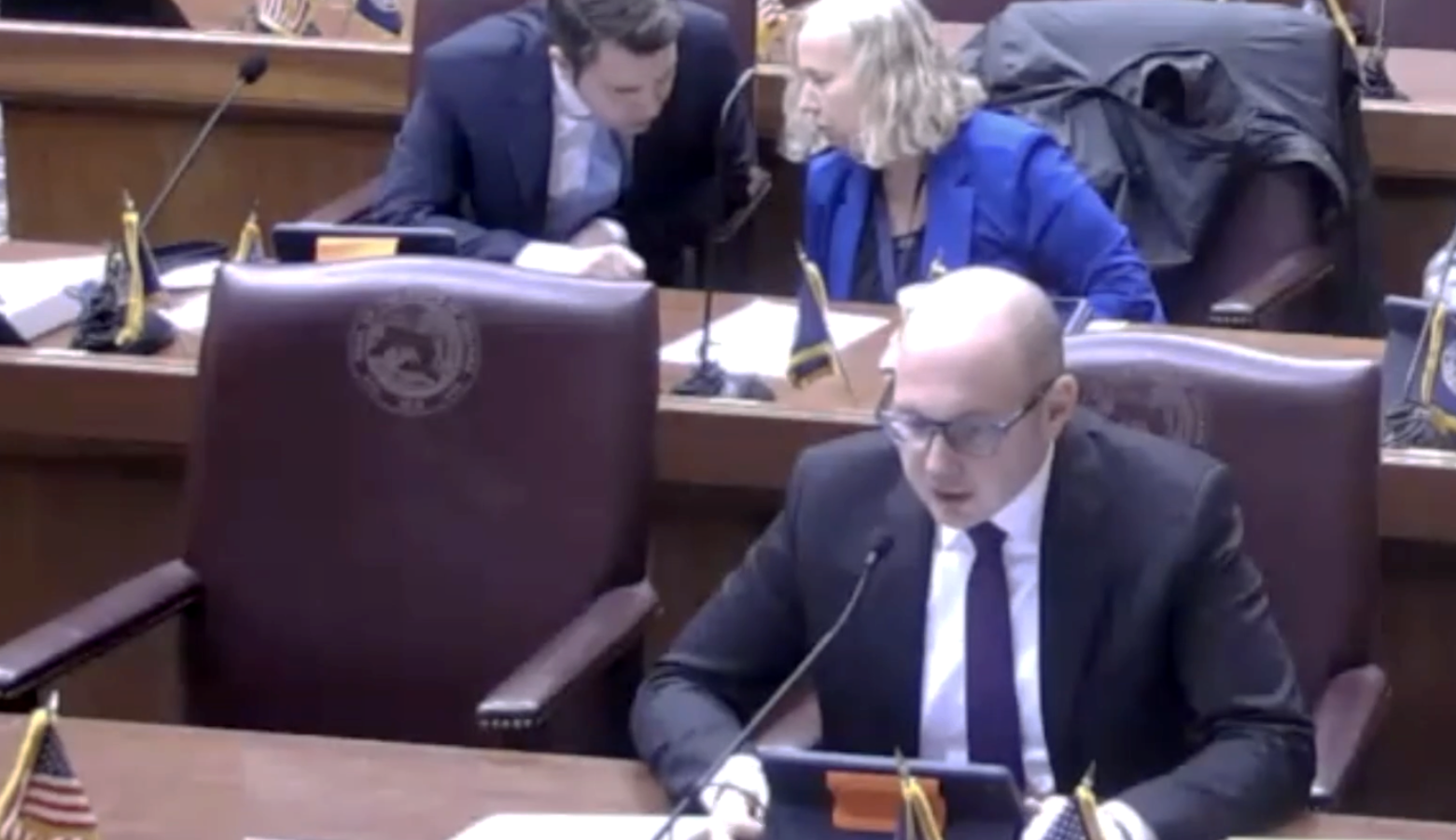 Jake Teshka sits on a red chair at a desk framed by small Indiana and United States flags. He is a White man, bald, wearing a suit and glasses. Two people whisper at the desk behind him.