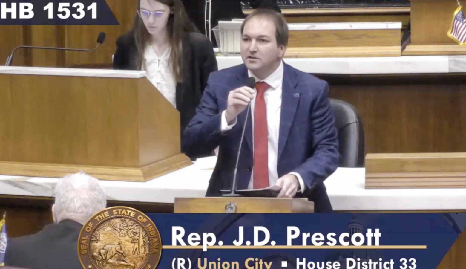 A screenshot of a livestream of the legislature shows J.D. Prescott speaking on the House floor. Prescott is a White man with brown hair. He is wearing a suit and tie.