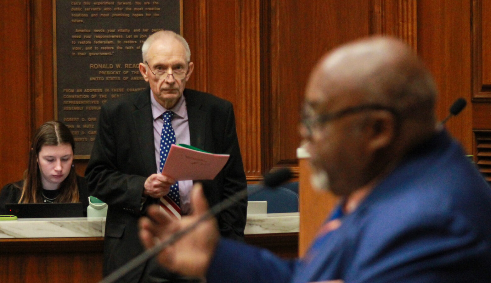 Jeff Thompson stands on the House floor, holding pieces of paper. Greg Porter stands at a lectern, speaking into a microphone. Thompson is a White man with white hair. He is wearing glasses and a suit and tie. Porter is a Black man, bald with a white goatee. He is wearing glasses and a suit and tie.