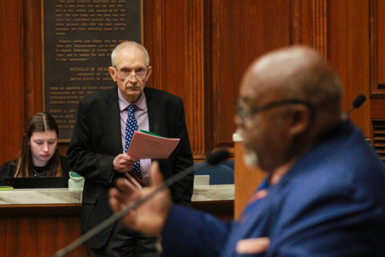 Jeff Thompson stands on the House floor, holding pieces of paper. Greg Porter stands at a lectern, speaking into a microphone. Thompson is a White man with white hair. He is wearing glasses and a suit and tie. Porter is a Black man, bald with a white goatee. He is wearing glasses and a suit and tie.