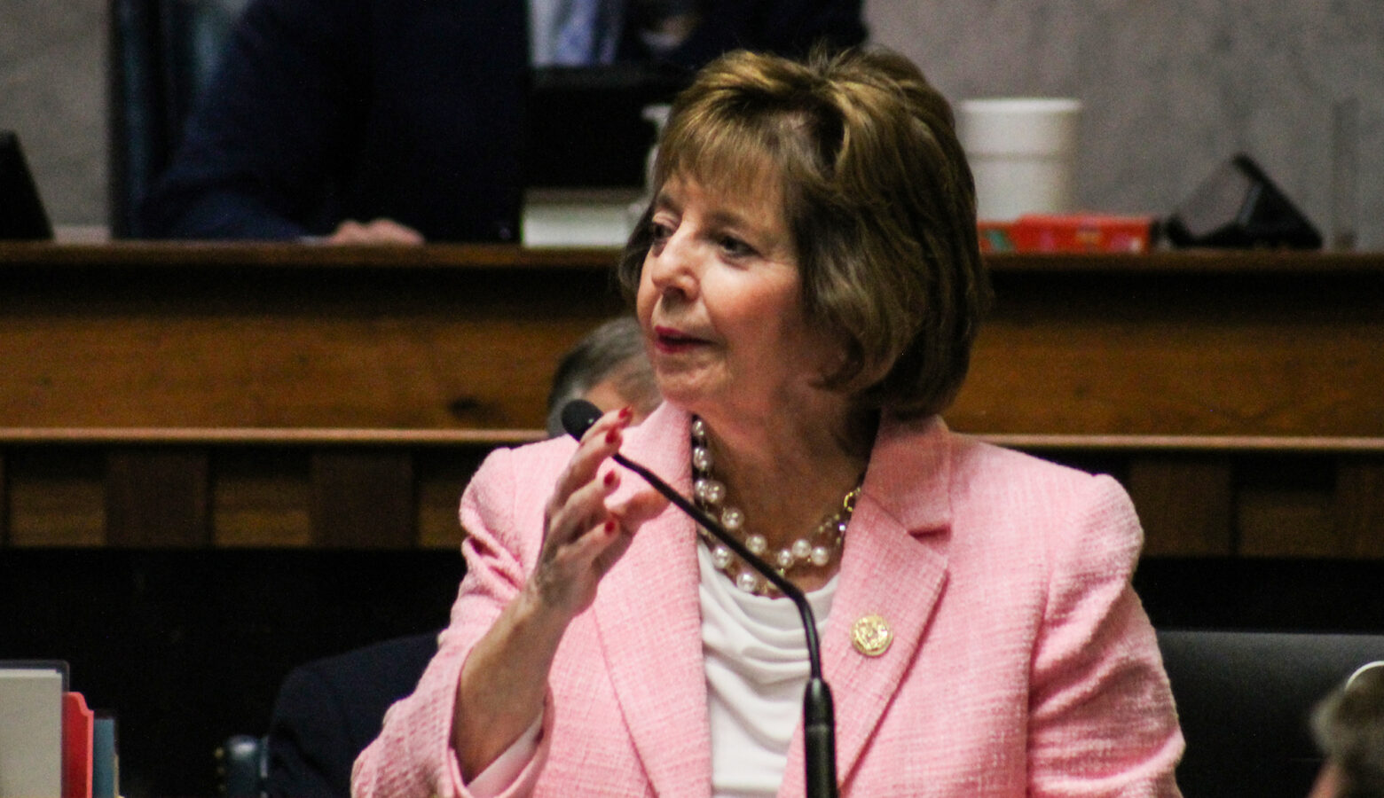 A woman in a light pink blazer wearing a gold pin and pearls turns her head and lifts her right hand as she speaks into a microphone in the Indiana Senate Chamber.