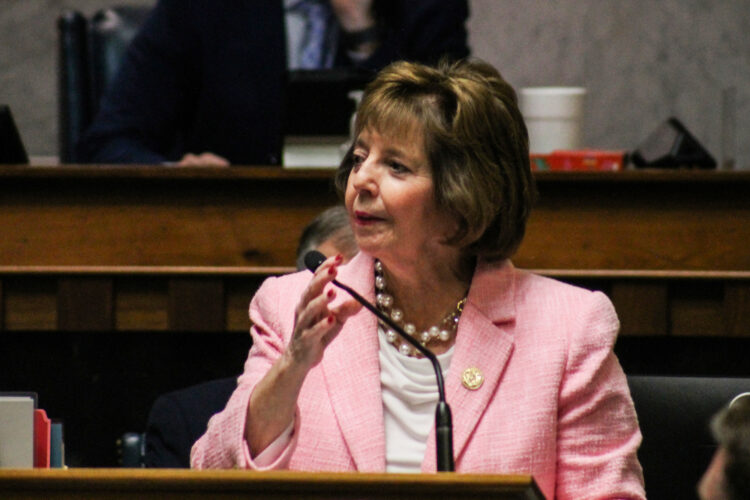 A woman in a light pink blazer wearing a gold pin and pearls turns her head and lifts her right hand as she speaks into a microphone in the Indiana Senate Chamber.