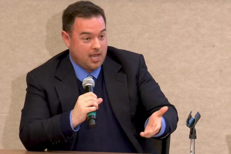 Luke Britt speaks into a microphone. He is a White man wearing a blue shirt, blue sweater, and black jacket.