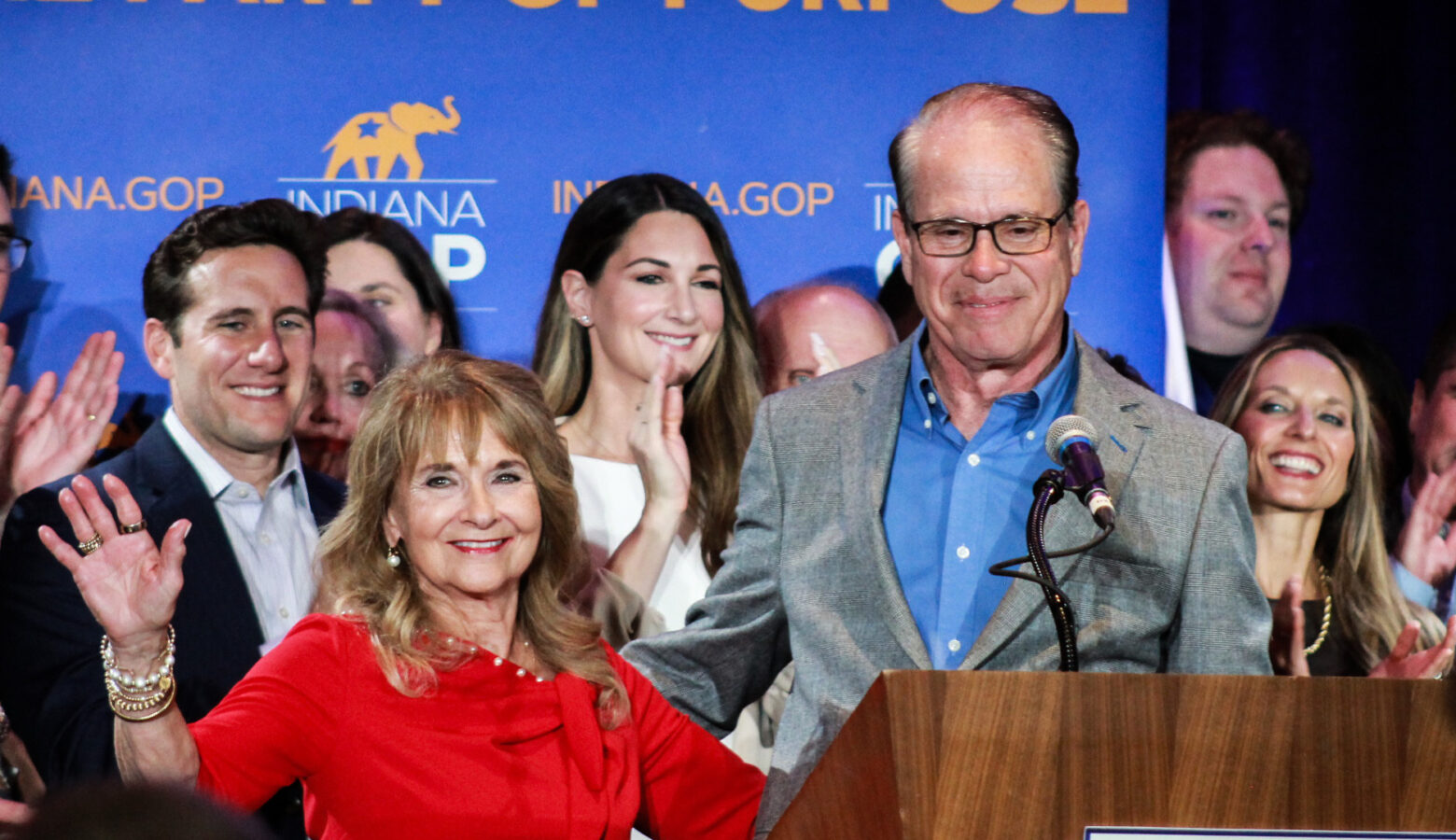 Maureen Braun is a White woman with light brown hair. She is wearing a red dress. Mike is a White man, balding with dark graying hair. He is wearing a gray suit jacket and blue shirt.