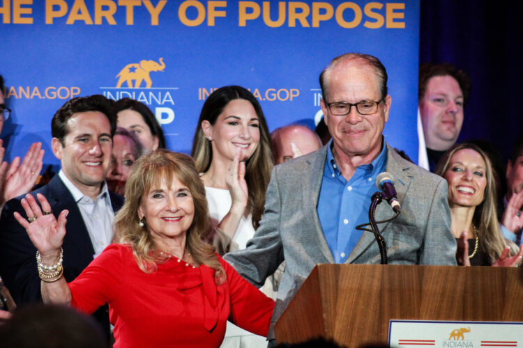 Maureen Braun is a White woman with light brown hair. She is wearing a red dress. Mike is a White man, balding with dark graying hair. He is wearing a gray suit jacket and blue shirt.