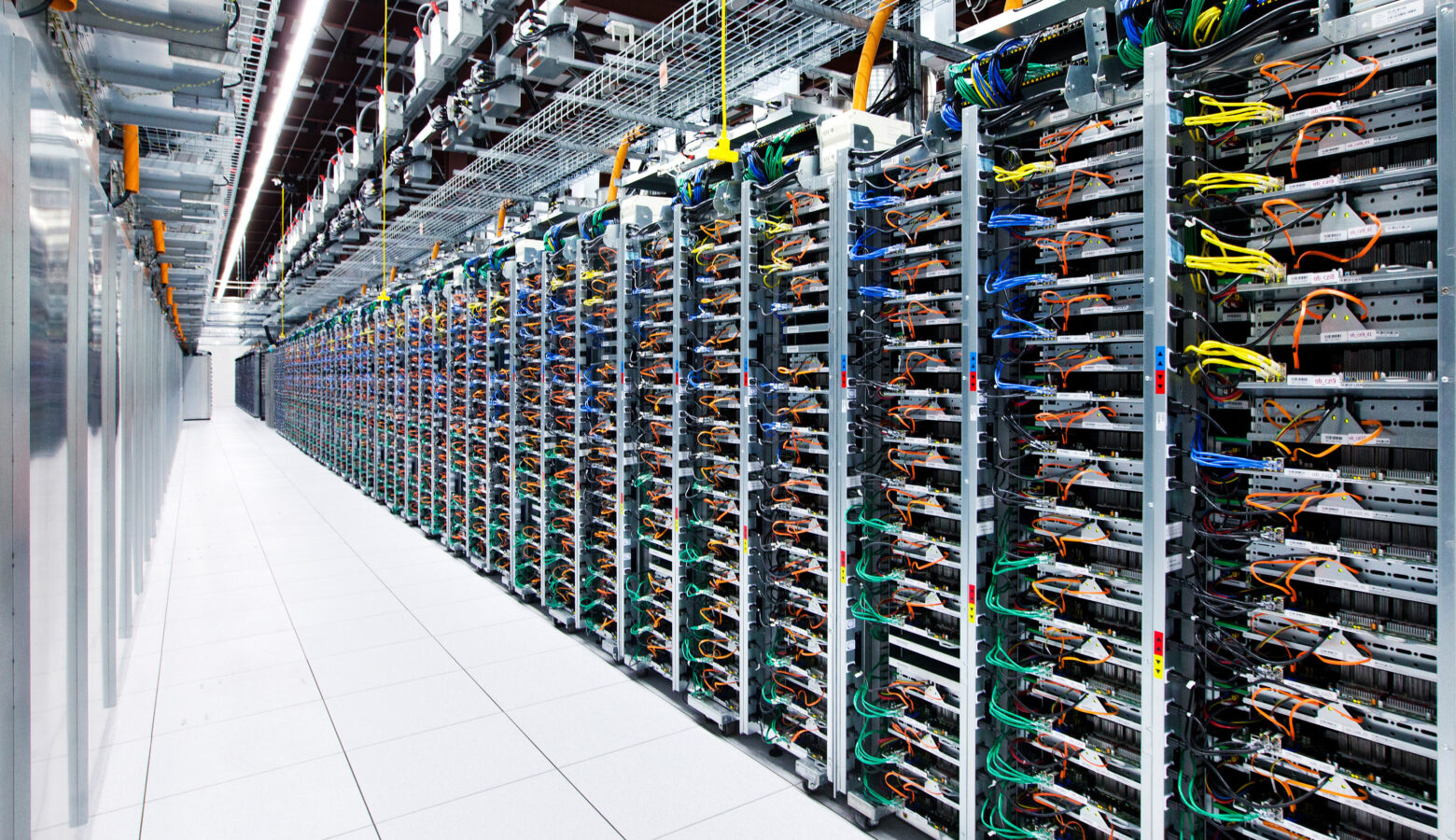 Server racks stretch down a long hallway. The tiled floor is white and wires poke neatly out of the metal racks.