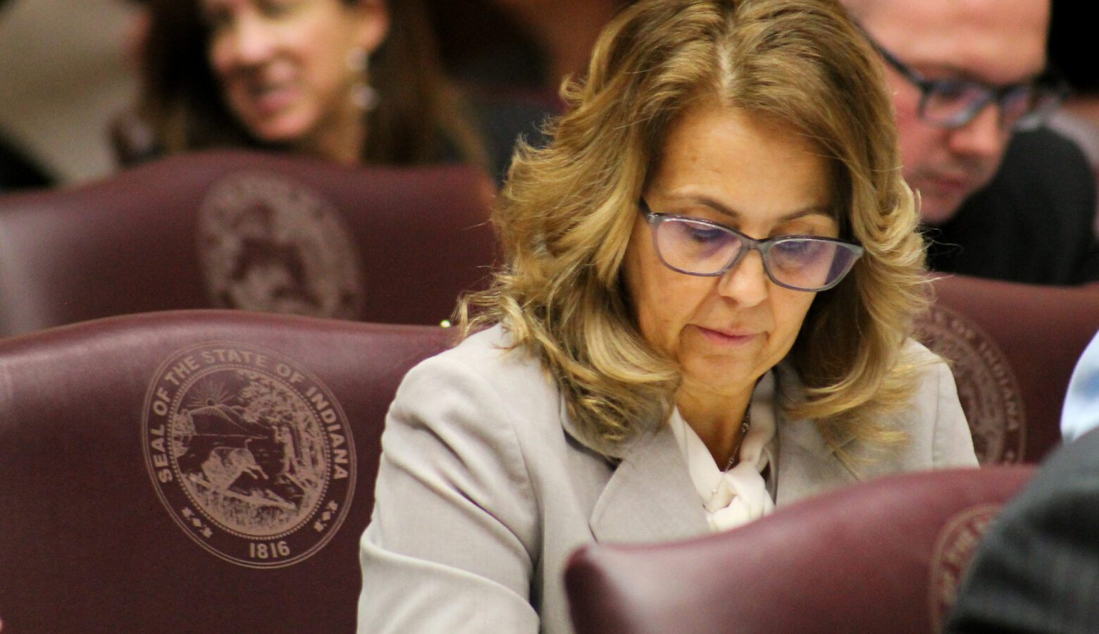 Representative Michelle Davis sits at her desk on the floor of the Indiana House. She is a White woman with glasses, wearing a grey suit.