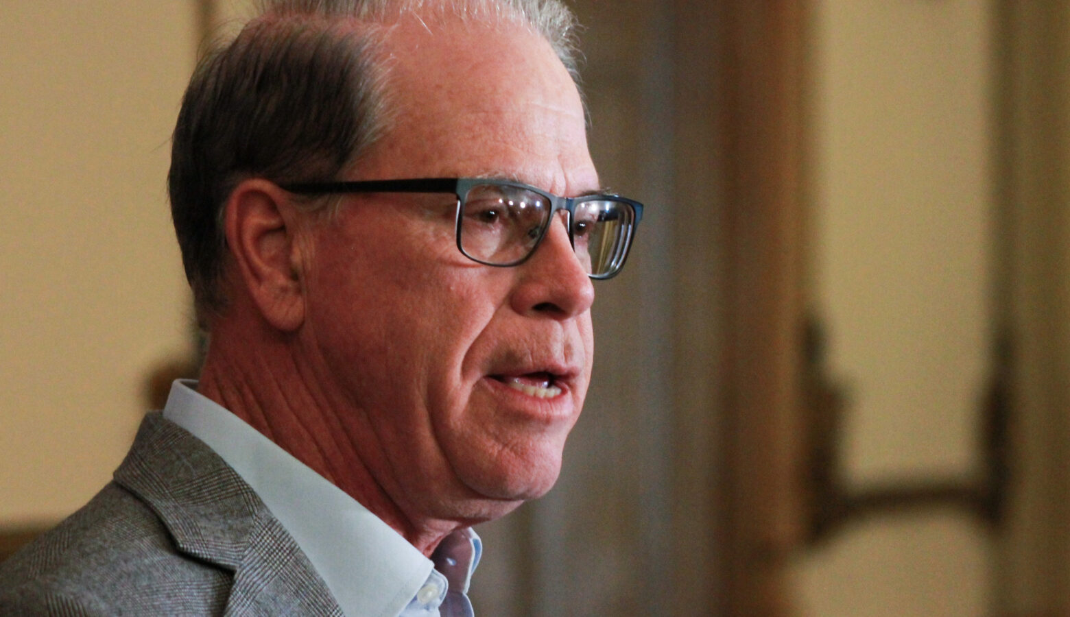 Mike Braun is a White man, balding with gray hair. He is wearing glasses and a gray blazer over a light blue shirt.