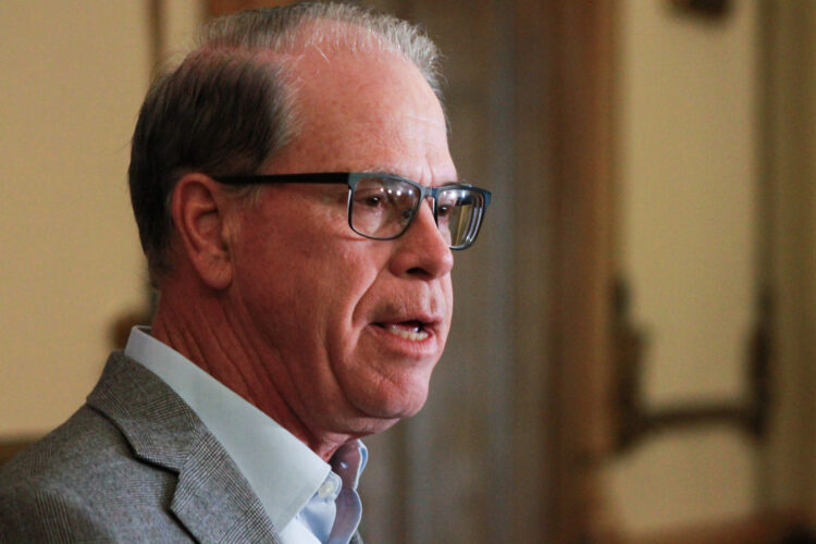 Mike Braun is a White man, balding with gray hair. He is wearing glasses and a gray blazer over a light blue shirt.