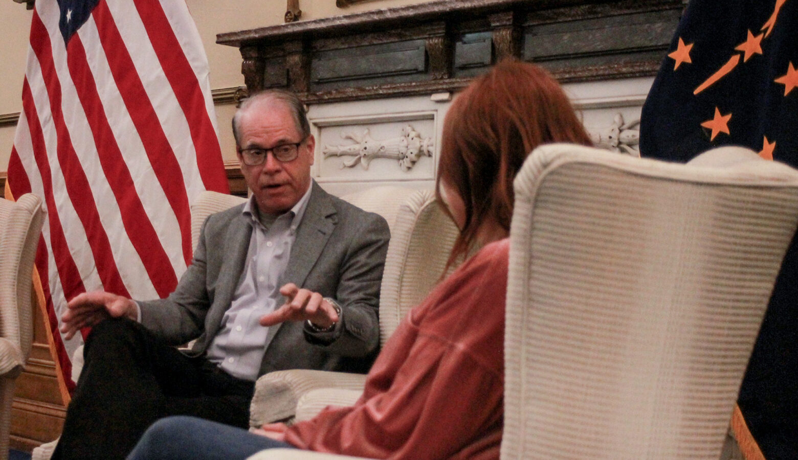 Mike Braun sits in a chair, talking with a woman who is facing away from camera. Braun is a White man, balding with gray hair. He is wearing glasses and a gray suit jacket.