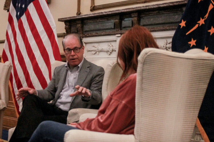 Mike Braun sits in a chair, talking with a woman who is facing away from camera. Braun is a White man, balding with gray hair. He is wearing glasses and a gray suit jacket.