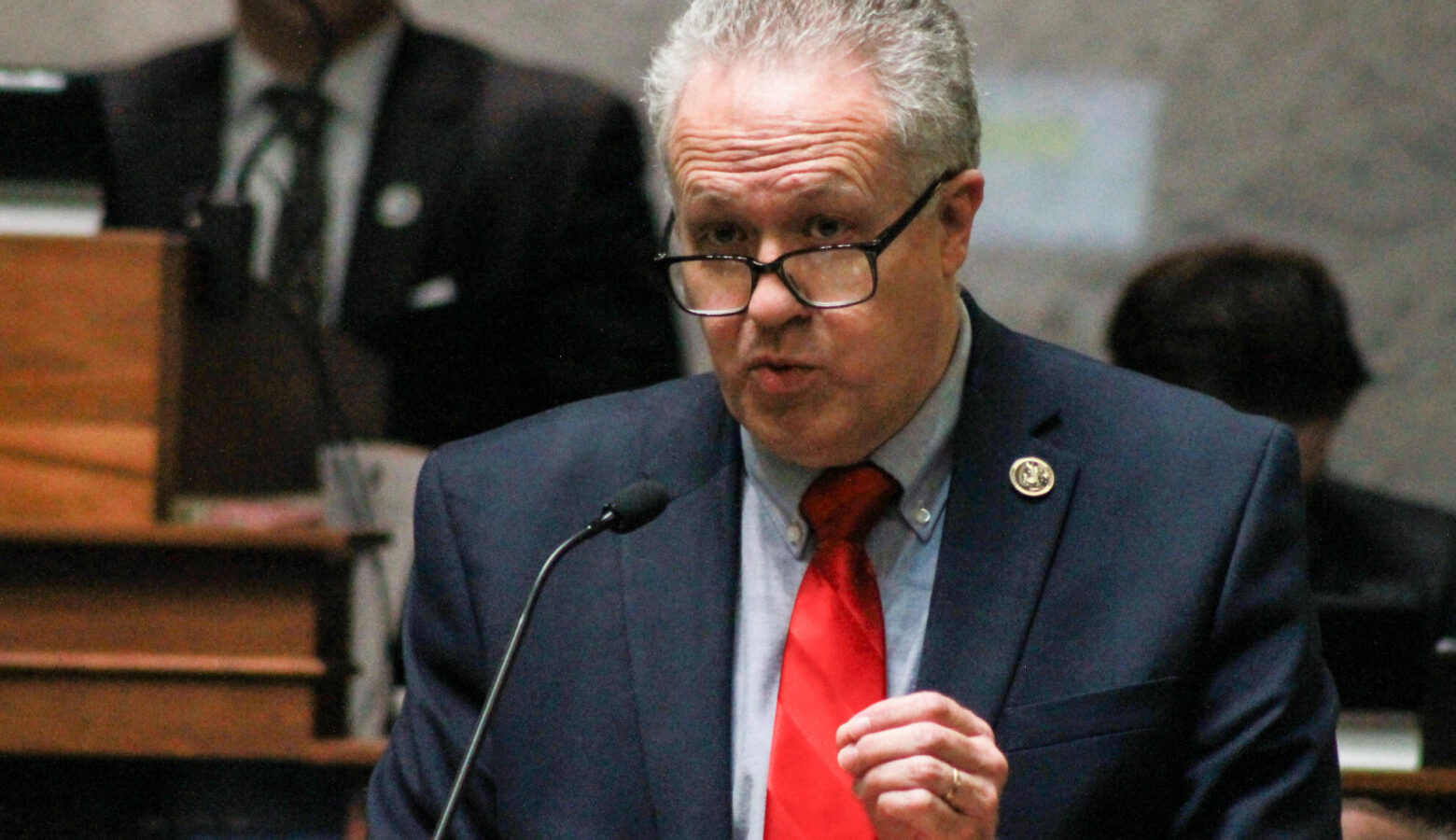 Mike Gaskill speaks on the Senate floor. Gaskill is a White man with white and gray hair. He is wearing glasses and a navy blue suit with red tie.