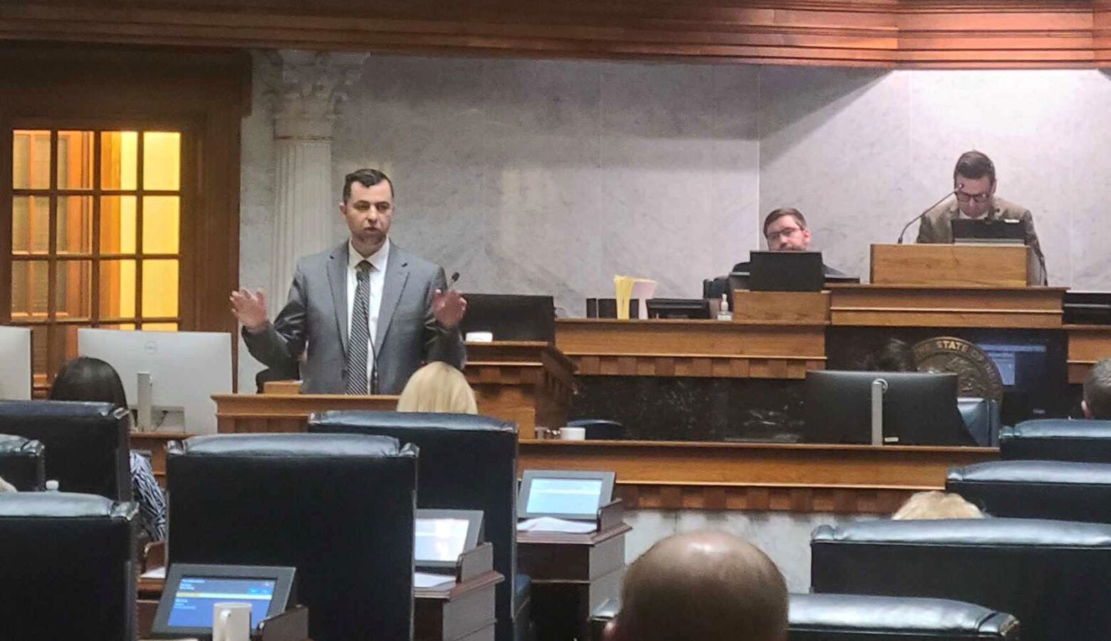 A man in a suit and tie stands behind a podium and lifts both hands in the air while addressing lawmakers in the Senate Chamber. The backs of senators' desks and chairs can be seen in front.