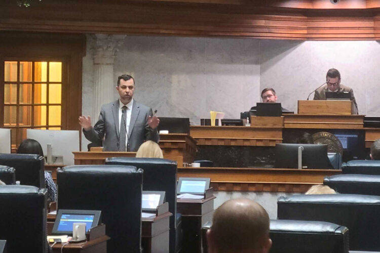 A man in a suit and tie stands behind a podium and lifts both hands in the air while addressing lawmakers in the Senate Chamber. The backs of senators' desks and chairs can be seen in front.