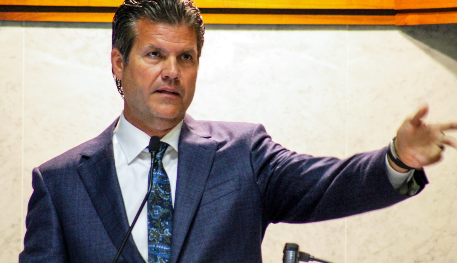 Senator Ryan Mishler wears a navy blue suit with a white shirt and a blue tie. He speaks at the front of the Senate chamber with his arm outstretched.