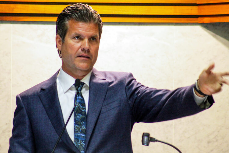 Senator Ryan Mishler wears a navy blue suit with a white shirt and a blue tie. He speaks at the front of the Senate chamber with his arm outstretched.