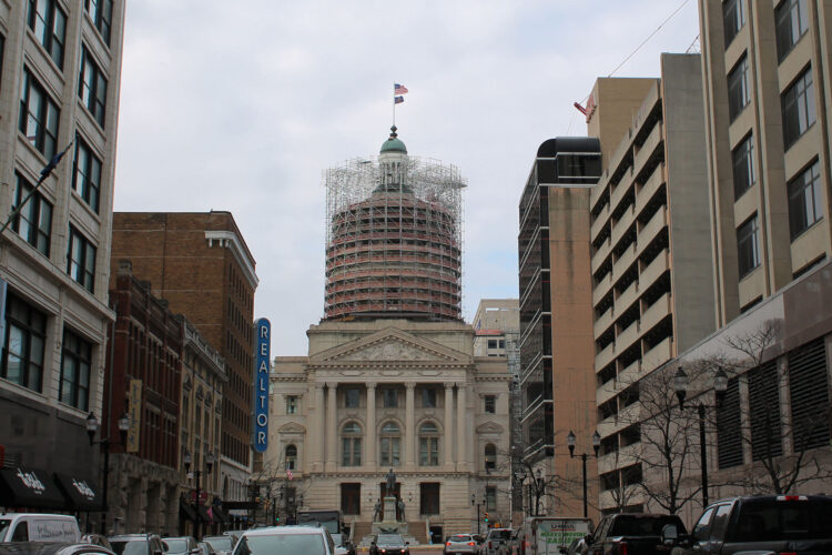 The eastern exterior of the Indiana Statehouse, framed between other buildings in downtown Indianapolis.