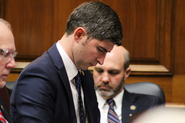 Tim Wesco listens to a speaker from his desk on the House floor. Wesco is a White man with dark hair. He is wearing a suit and tie.