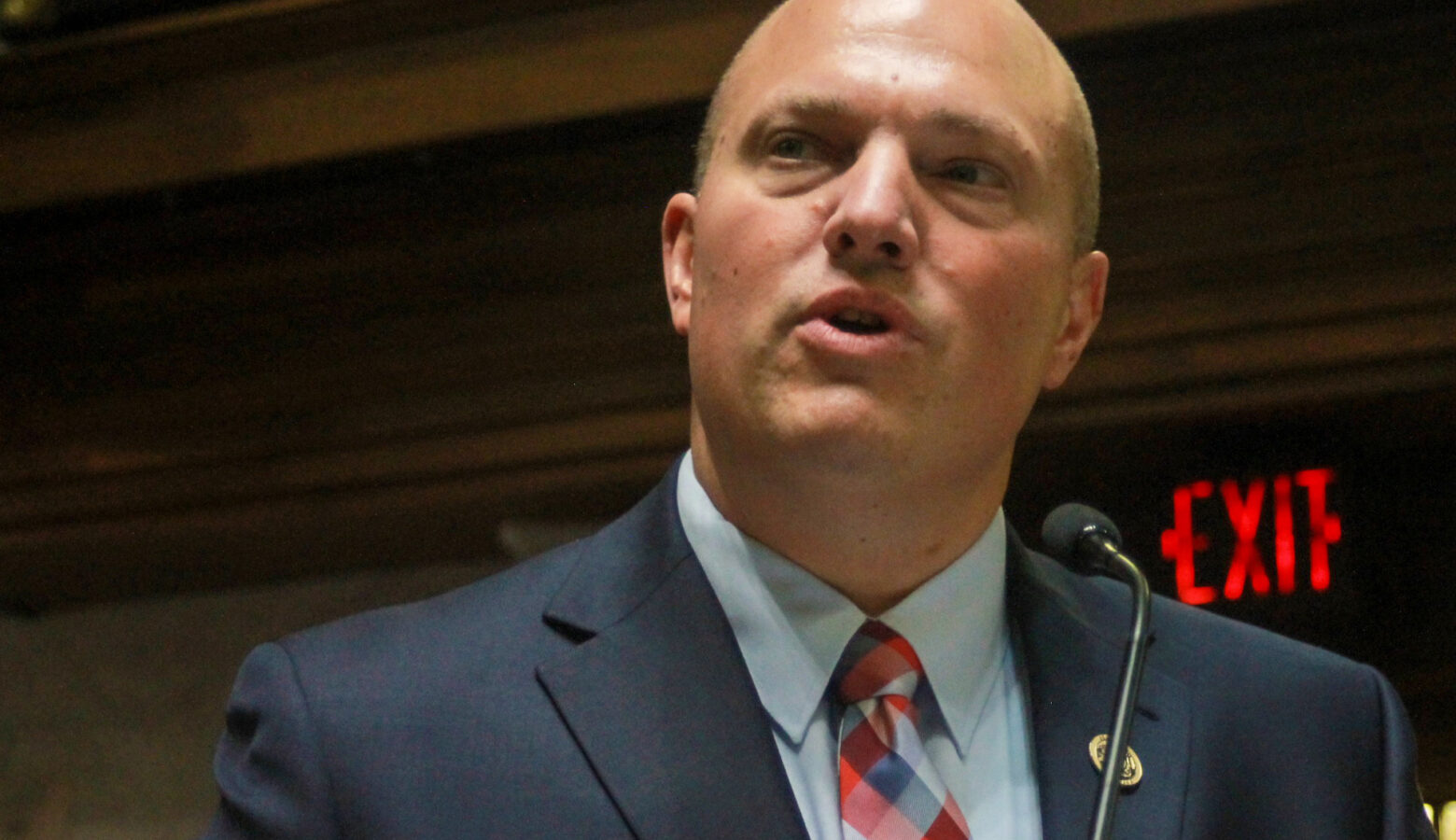 Senator Tyler Johnson wears a navy blue suit with a light blue shirt and a red, blue and light blue plaid tie. He is a bald White man.