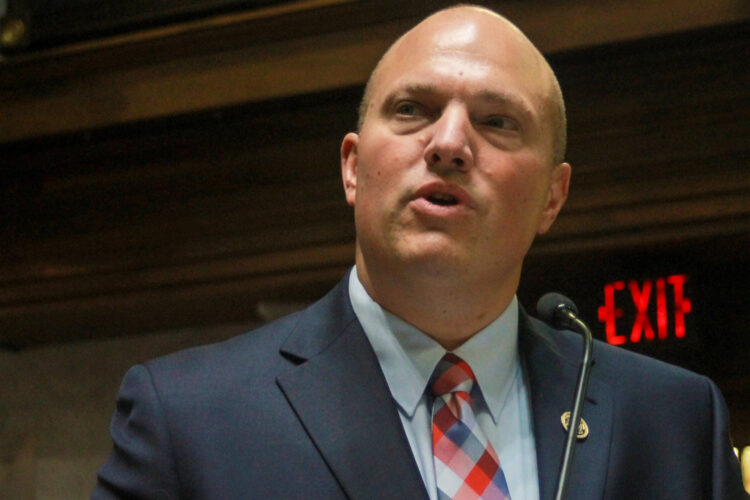 Senator Tyler Johnson wears a navy blue suit with a light blue shirt and a red, blue and light blue plaid tie. He is a bald White man.