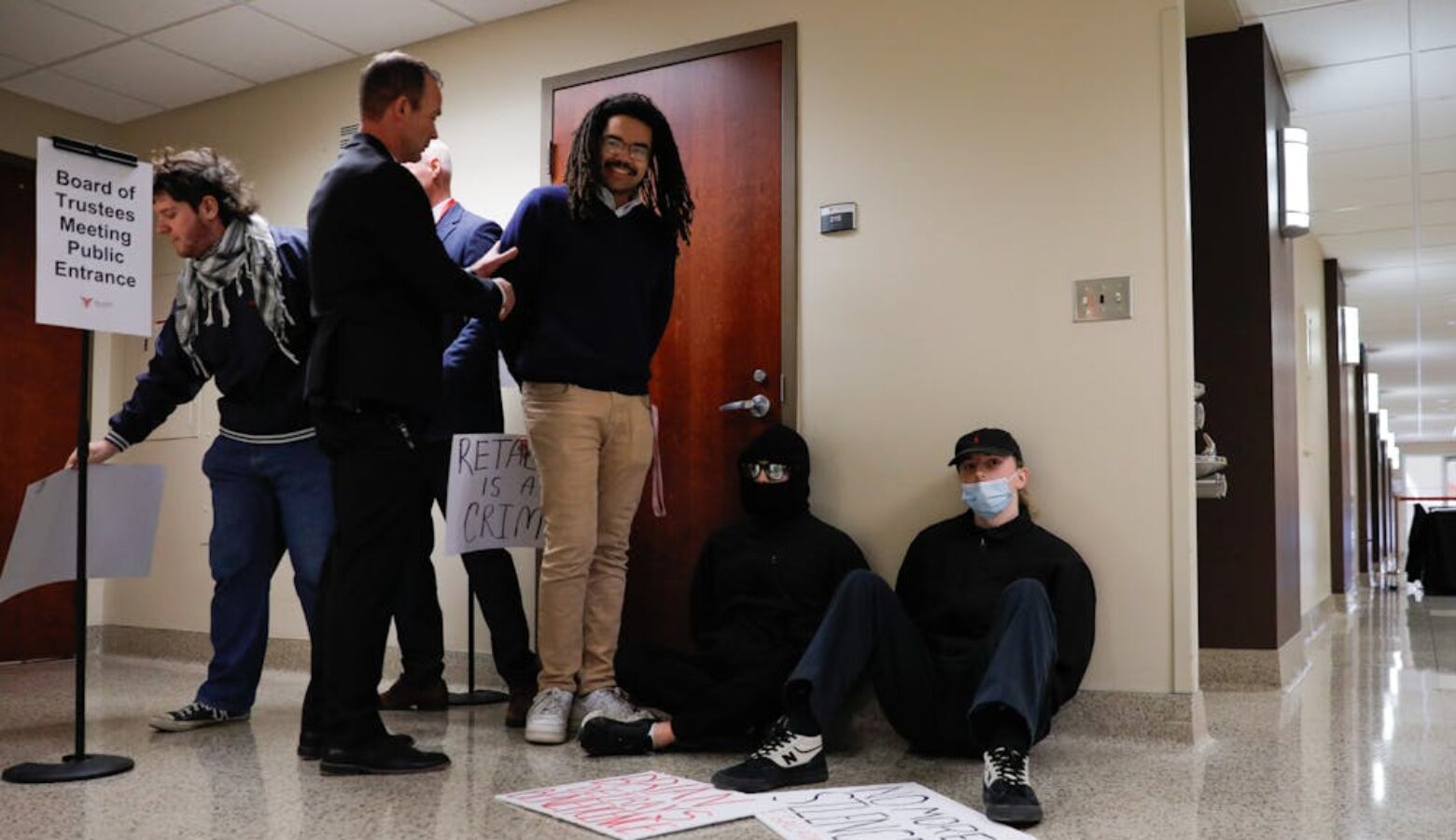 One man detains another man in a hallway, while two others sit against a wall. All have their hands held behind their back.