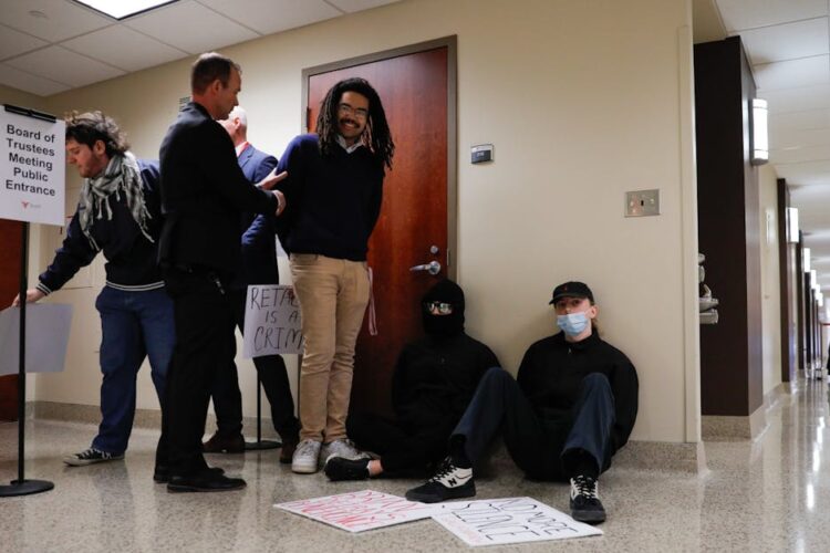 One man detains another man in a hallway, while two others sit against a wall. All have their hands held behind their back.