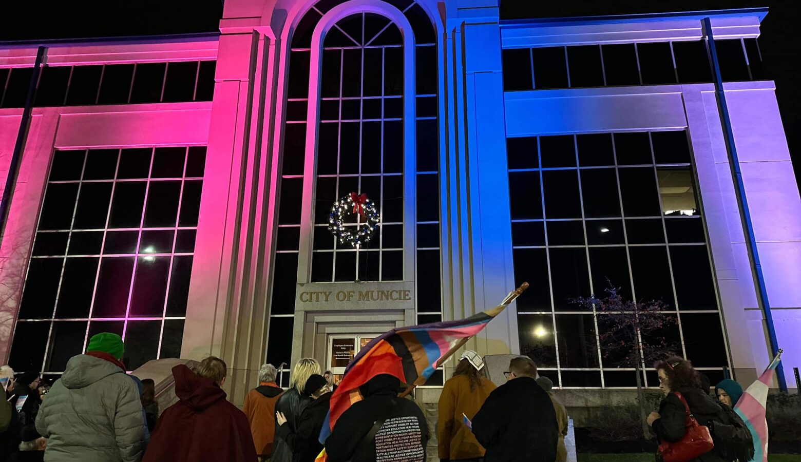 The Muncie City Hall is lit with pink and blue lights. There are people standing in front, one carrying a pride flag.