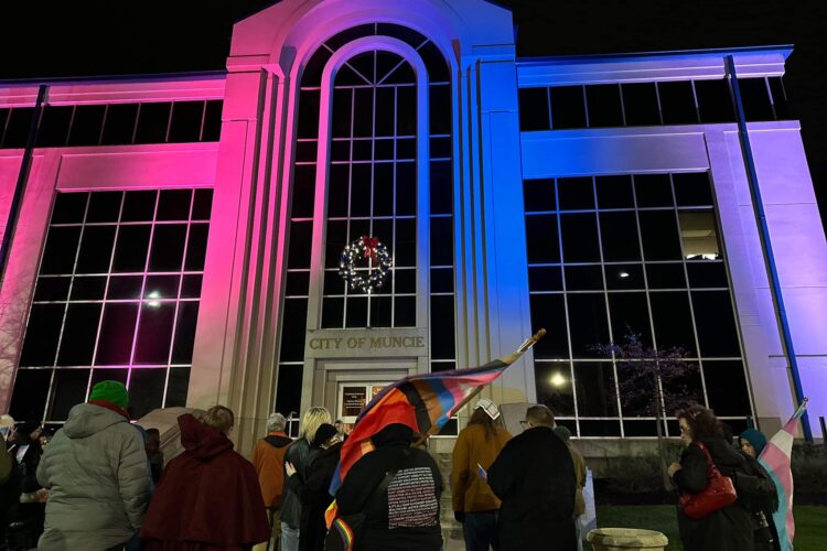 The Muncie City Hall is lit with pink and blue lights. There are people standing in front, one carrying a pride flag.