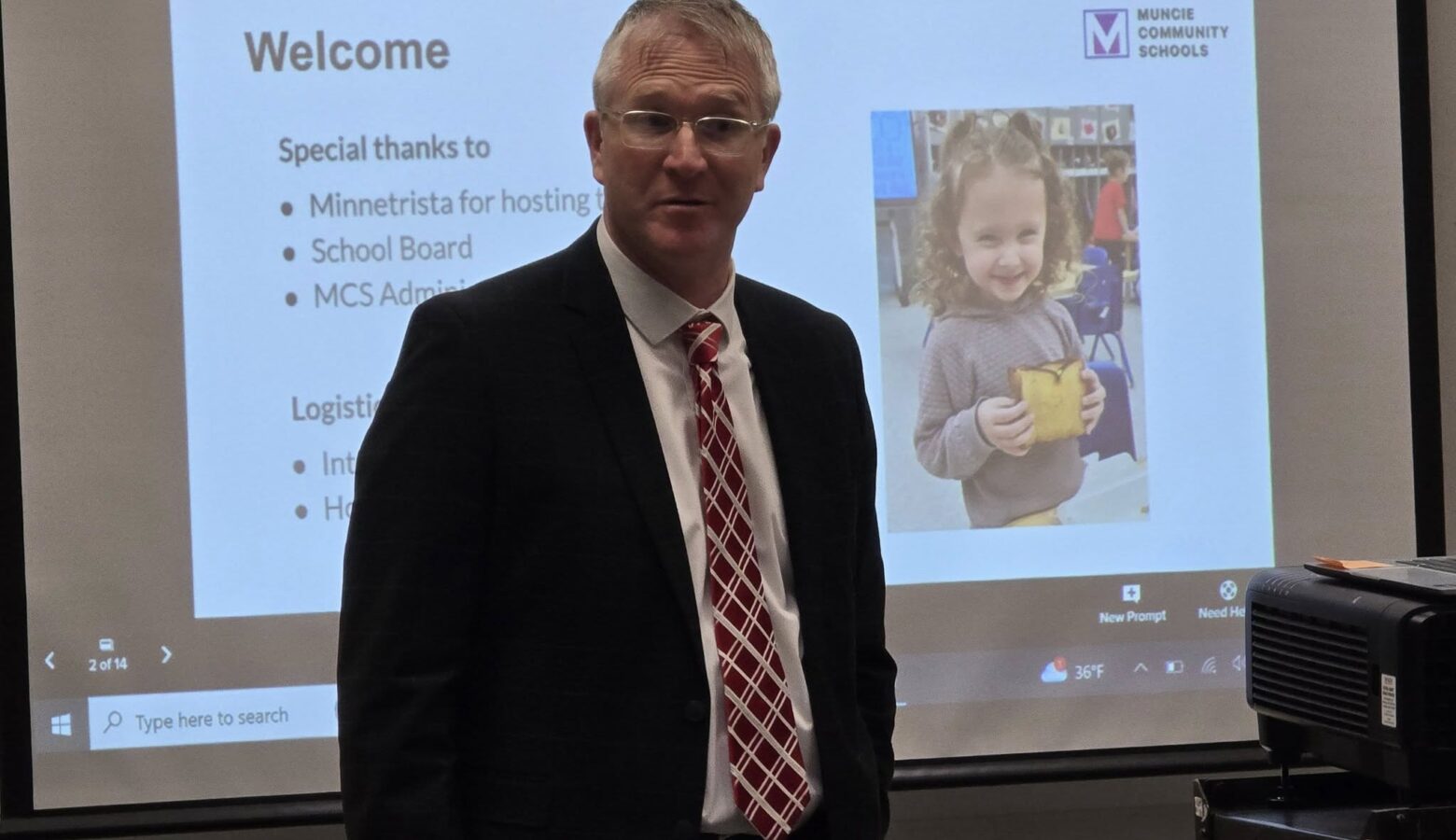 Chuck Reynolds stands in front of a presentation screen. He is a White man with glasses, wearing a black suit and red tie.