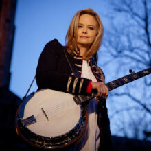 A woman with blonde hair wearing a banjo looks down at the camera.