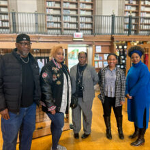 Five people stand in front of racks of books and art displays in a library.