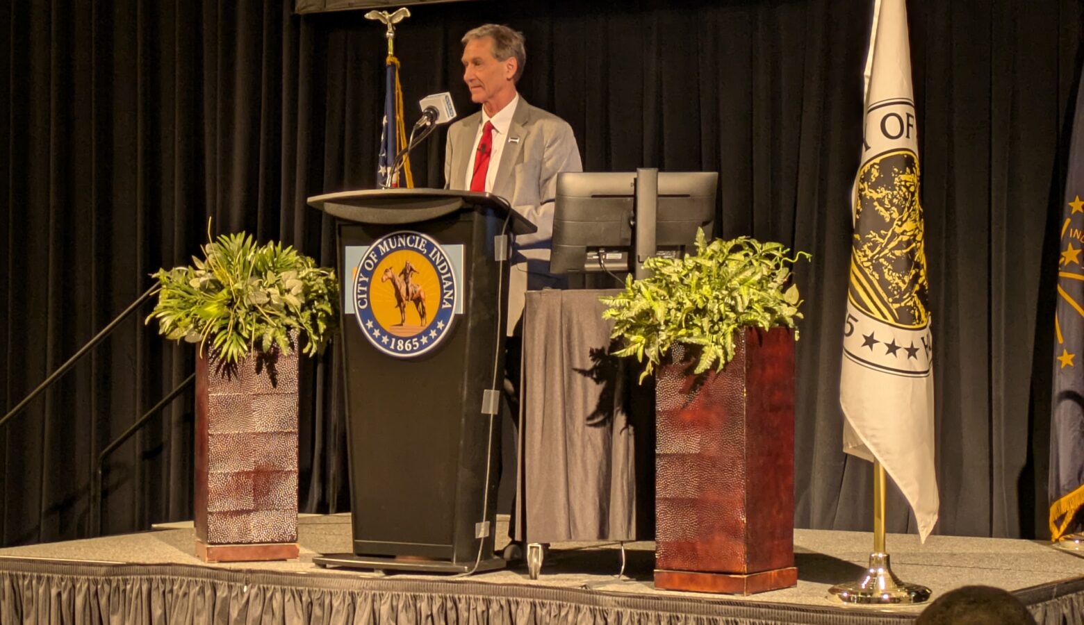 Muncie Mayor Dan Ridenour stands on a stage at a podium, surrounded by plants and flags. He is a White man wearing a gray suit and red tie.