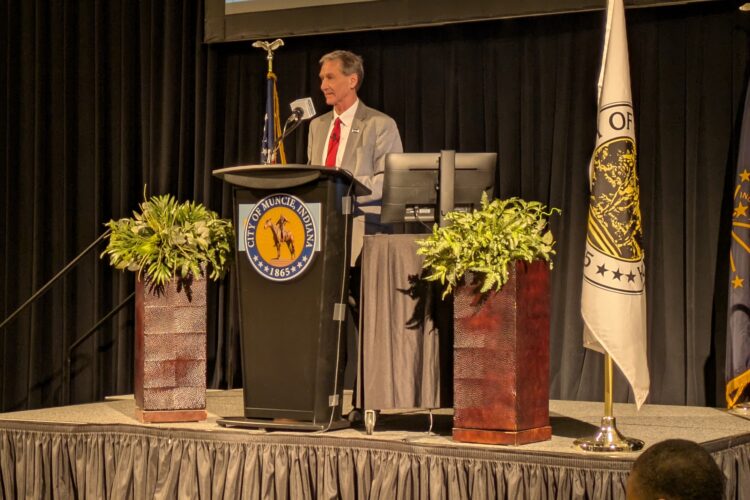 Muncie Mayor Dan Ridenour stands on a stage at a podium, surrounded by plants and flags. He is a White man wearing a gray suit and red tie.